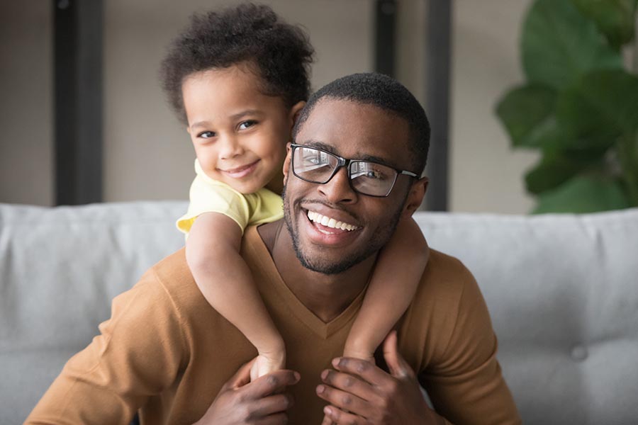 Personal Insurance - Dad Smiles as His Toddler Son Wraps His Arms Over Dad's Shoulders While Perched on Their Couch